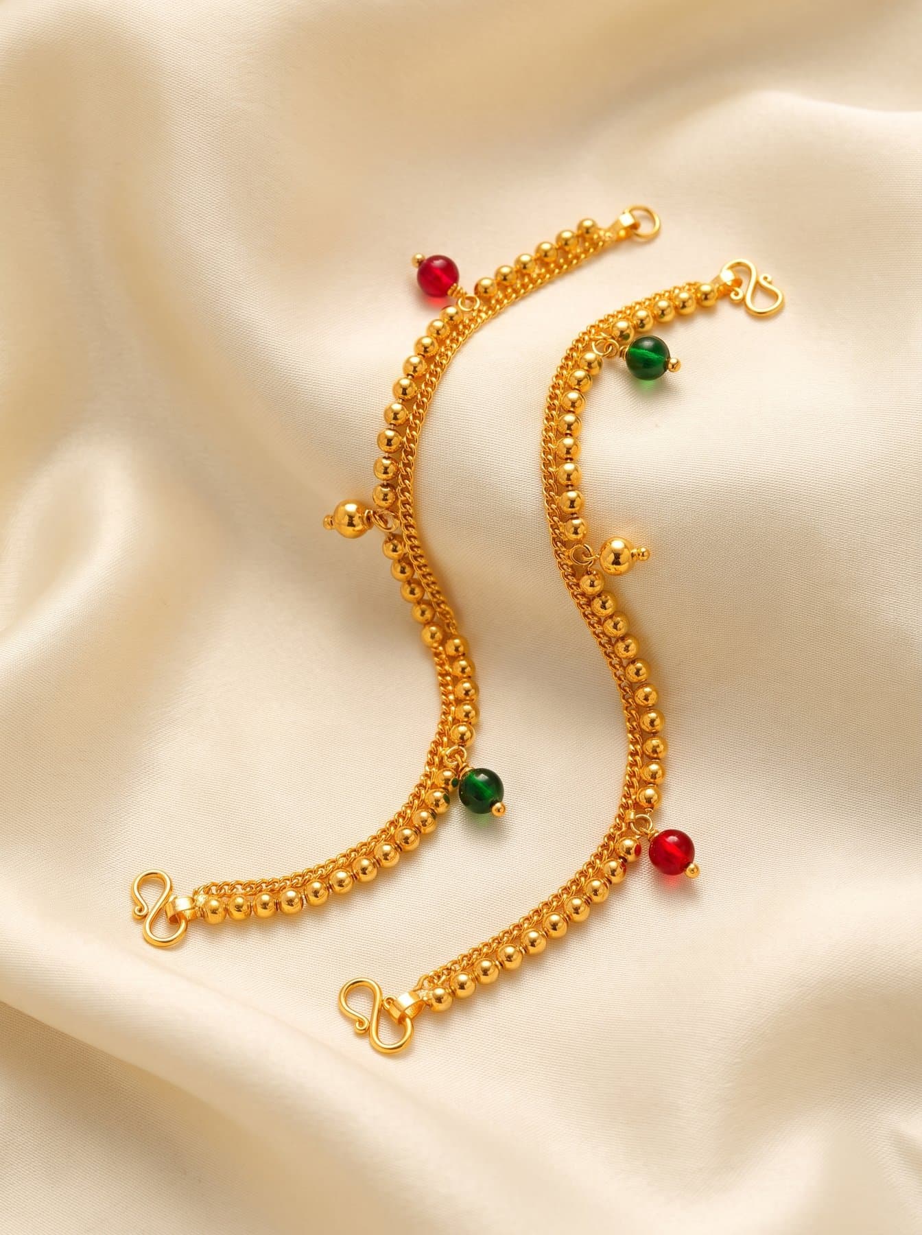 Pair of gold anklets with ruby-red and green beads laid on ivory silk