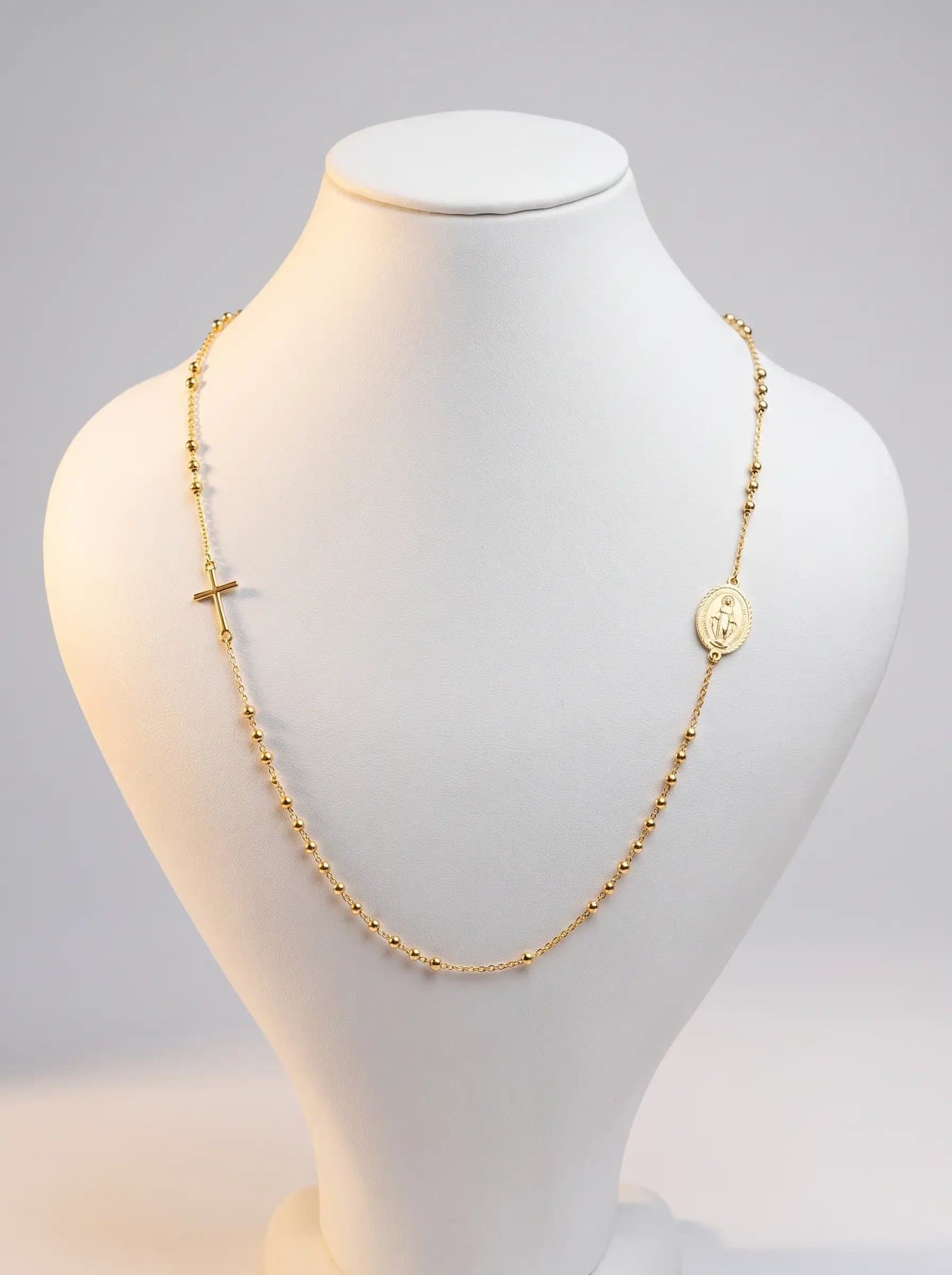 Gold beaded rosary chain with a sideways cross and Marian medallion on a white display bust